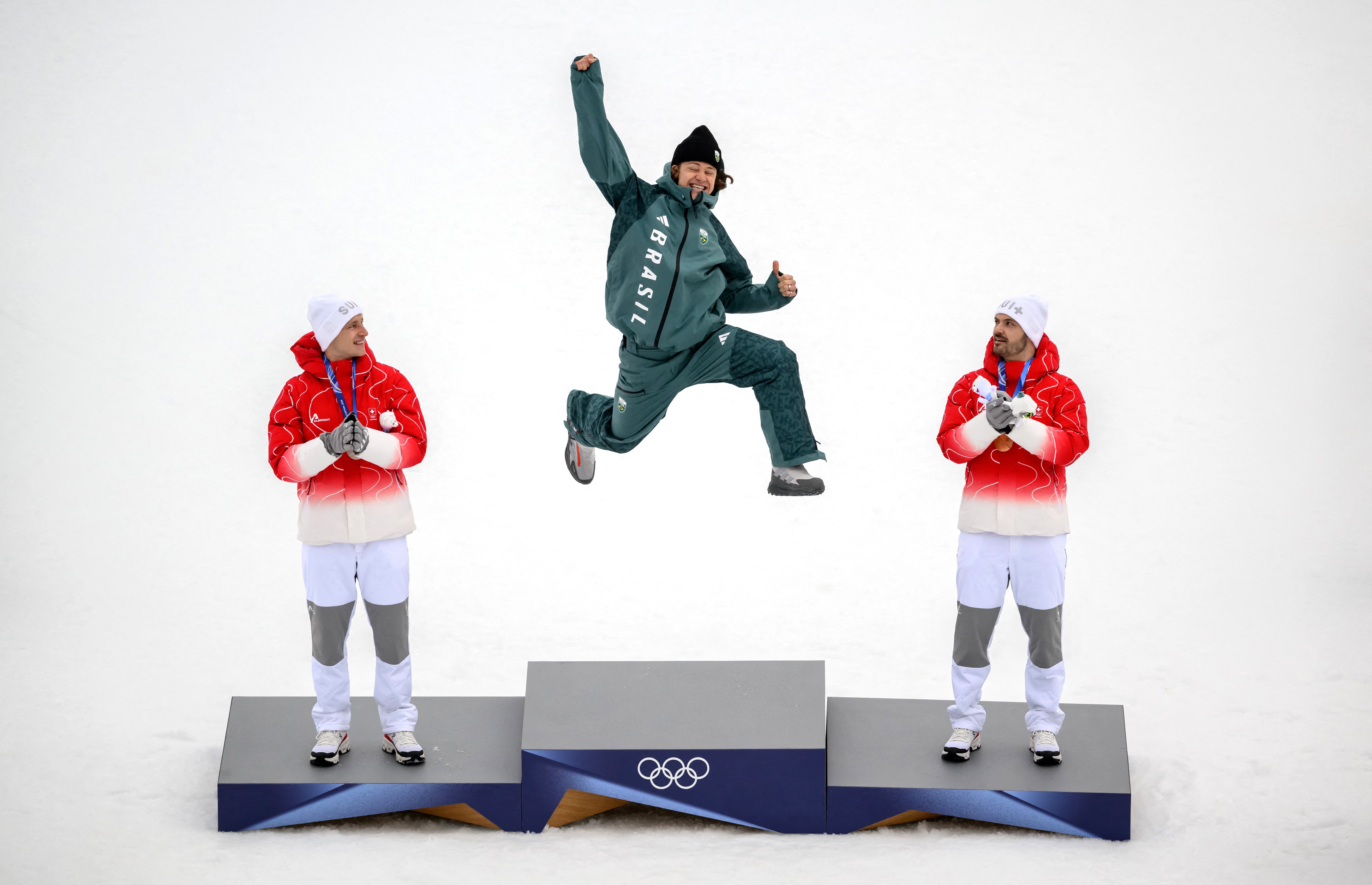 A skier jumps up, fist held high, smiling, as he climbs atop a podium to receive a gold medal, flanked by the bronze and silver winners.