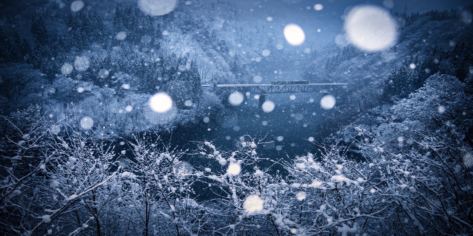 A wintry scene, with a train passing over a river bridge, seen through falling snow.