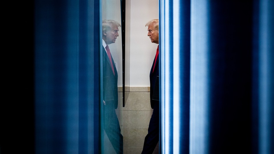 A color photograph of Donald Trump in profile, wearing a suit and red tie and looking at his reflection