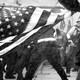 A black-and-white photo of an American flag held sideways in front of a row of cops facing off against men in tactical vests and MAGA sweatshirts.