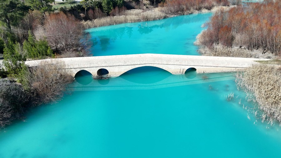 An aerial view of a bright-turquoise river flowing beneath a small stone bridge