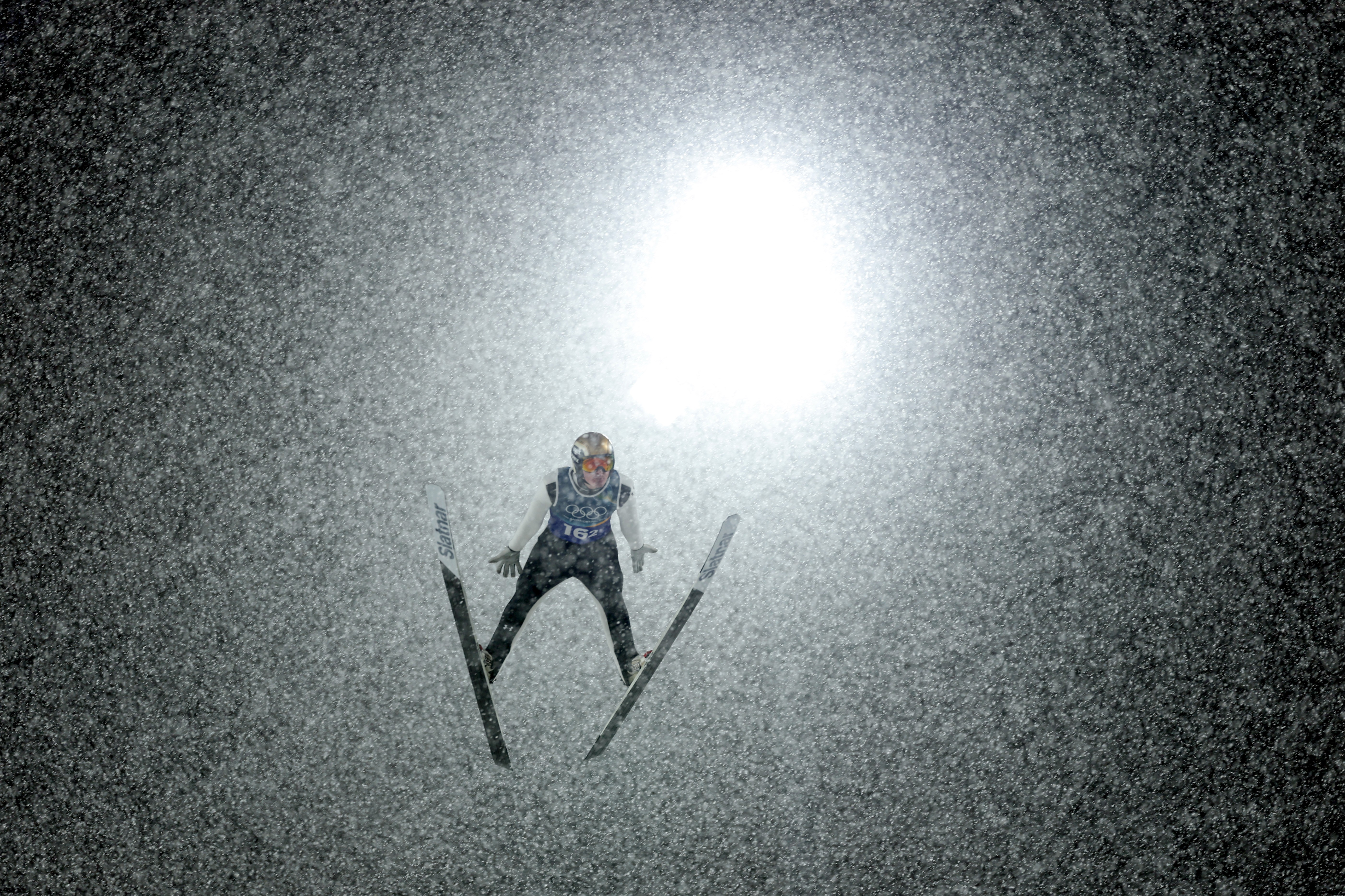 A ski jumper flies through a flurry of snowflakes at night, backlit by a bright light.
