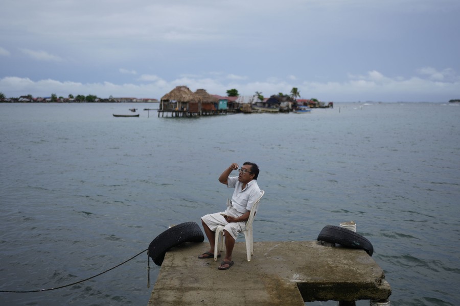 A person with a cellphone sits on a plastic chair on a small concrete dock. A tiny island with houses on stilts is visible in the background.