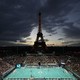 A view of a small football stadium at night, filled with spectators, beneath the Eiffel Tower
