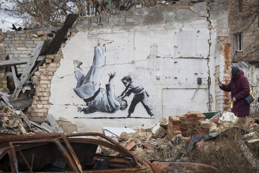 A woman takes a picture of artwork made by Banksy on a destroyed building wall, depicting a child throwing an adult with a judo move.