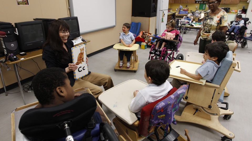 A woman holds a board with a cat drawing and the letter "C." A semicircle of students with disabilities surrounds her