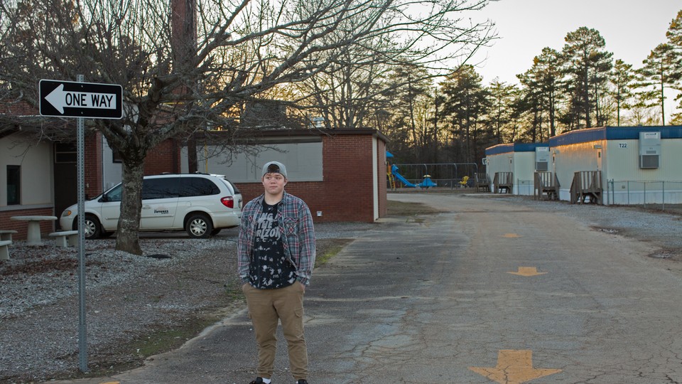 A 16-year-old boy wearing a backward baseball cap, flannel shirt, and khakis stands in front of trailers and a "one way" sign with his hands in his pockets.