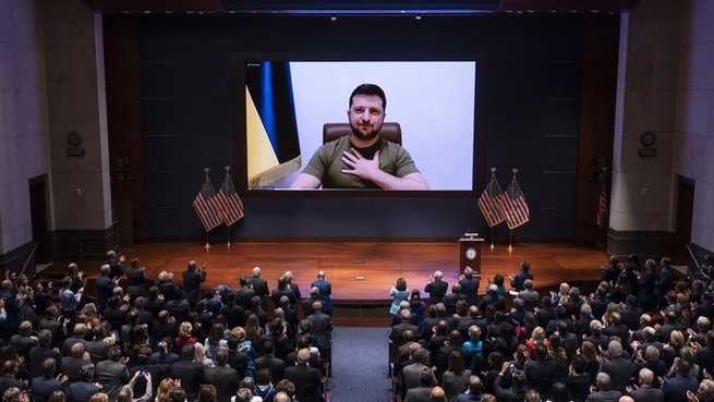 Members of Congress stand in front of a large screen that shows the president of Ukraine putting his hand over his heart.