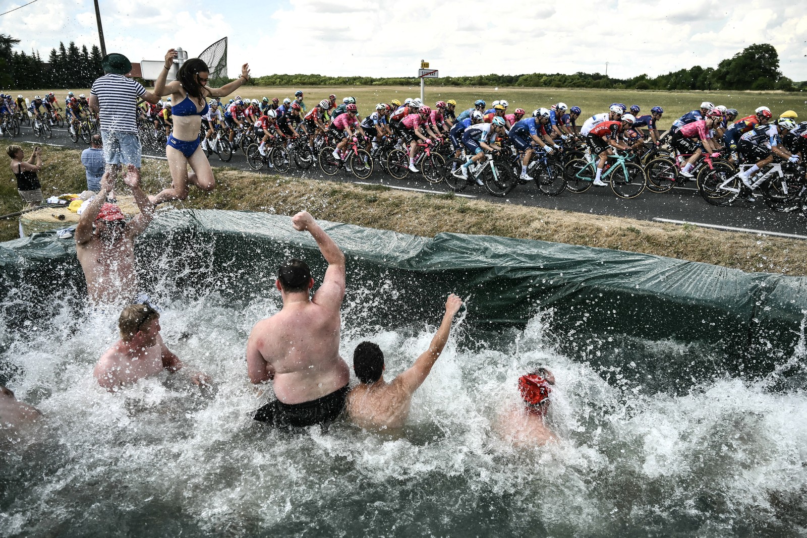 Spectators jump and splash in a makeshift swimming pool set up along a race route as a pack of riders cycles past.