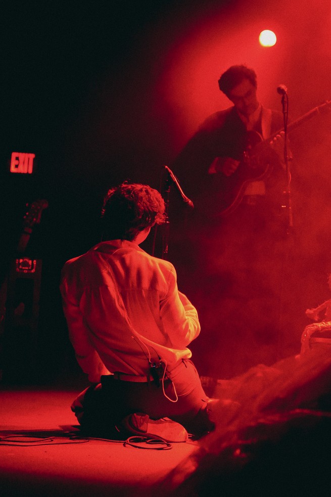 Singer on his knees in front of guitarist all bathed in a red glow