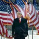 Donald Trump standing outside, behind clear barriers and in front of a row of billowing American flags