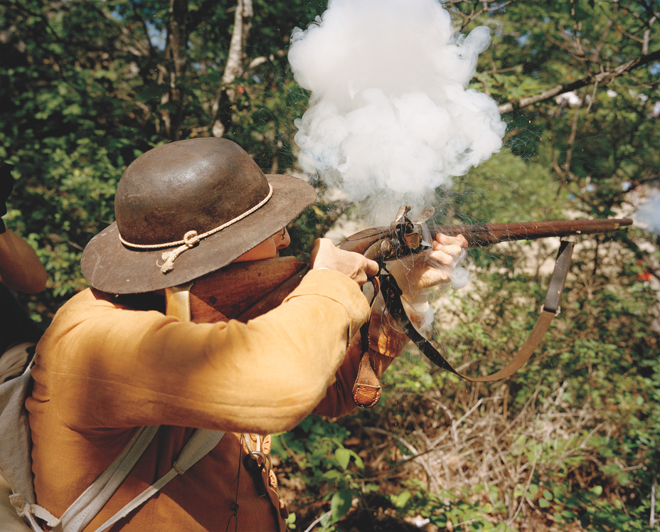 photo of a reenactor wearing a leather brimmed hat with rope tie and tan jacket kneeling and firing a musket through trees, with a cloud of smoke 