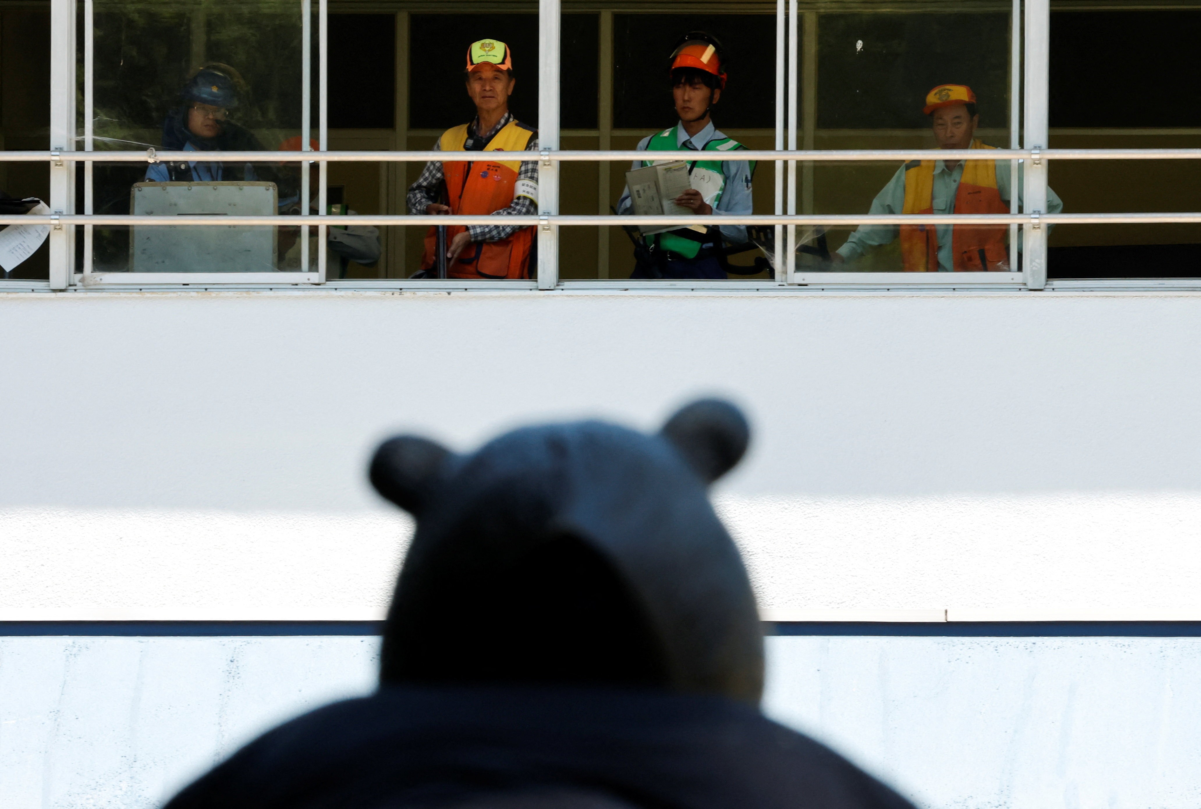 A view of several hunters and government workers in a bank of windows, looking outside, seen over the head of someone in a bear mask, in the foreground