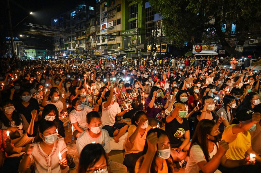 A city street is filled with people who are seated, holding candles and raising their fists.