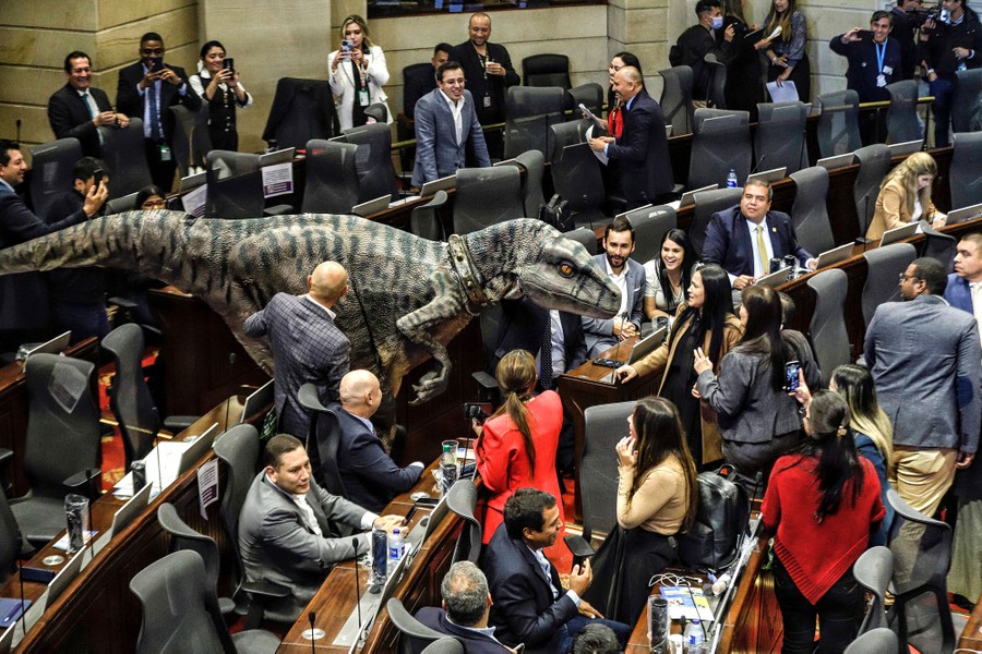 A person in a dinosaur costume walks among lawmakers in a large room.