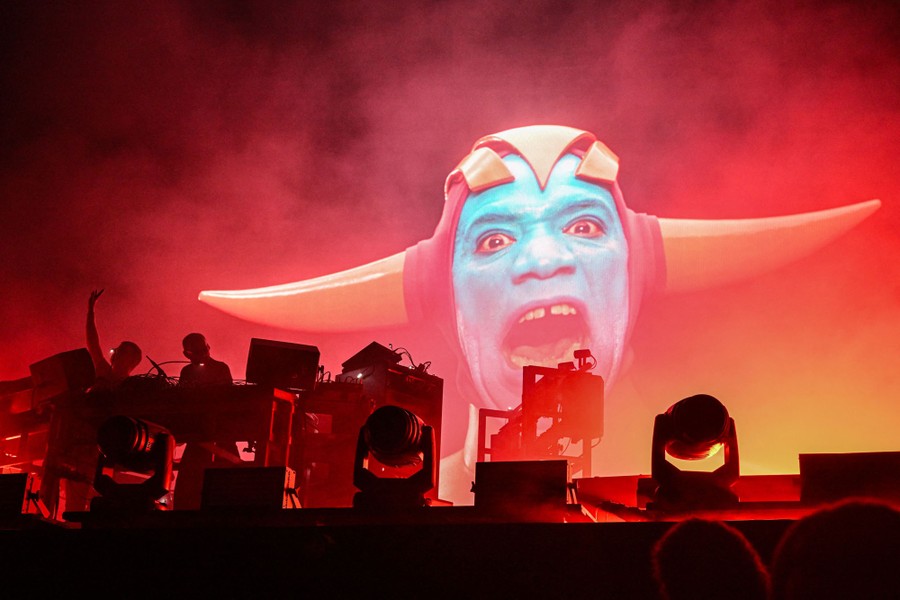 Musicians perform on a stage in front of a screen projection of a blue-faced person wearing a horned costume.