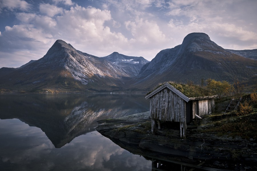 A partly overgrown boathouse stands in front of a mountain vista.