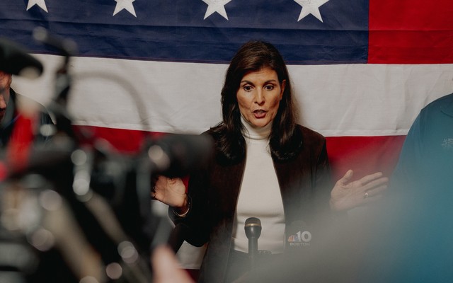 Photo of Nikki Haley speaking at a microphone, hands apart, with press before her and an American flag behind her