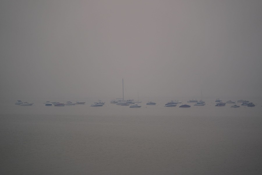 Many boats are seeing floating in a lake beneath a very smoky sky.