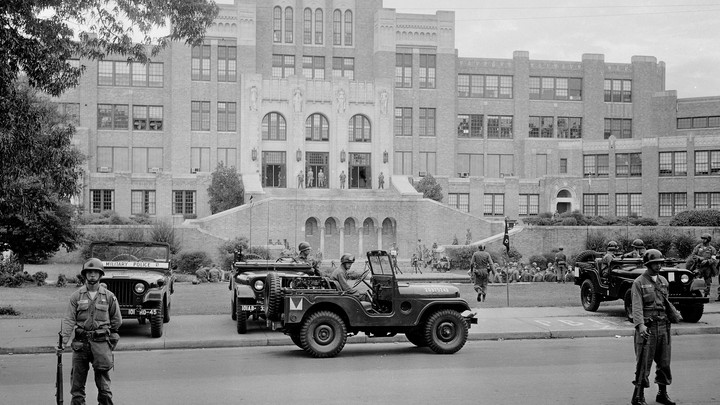 This black and white photo shows men in combat attire standing guard outside a large brick school building. Three military-esque Jeeps are in the photo.