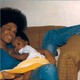 A woman sitting in a brown chair with her leg thrown over the arm holds a baby while looking at the camera.