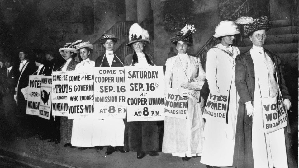 A black and white photo of women in dresses and feathered hats. They hold signs advertising an upcoming rally for women's right to vote.