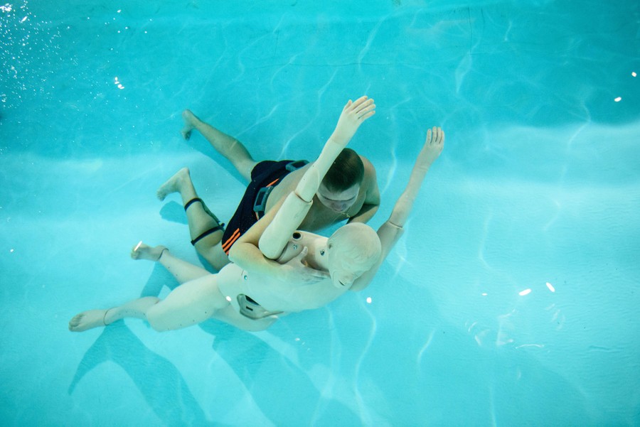 A person carries a mannequin underwater in a pool.