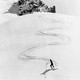 A black and white photograph of a skier coming down a steep alpine with a zig zag-pattern in the snow behind them