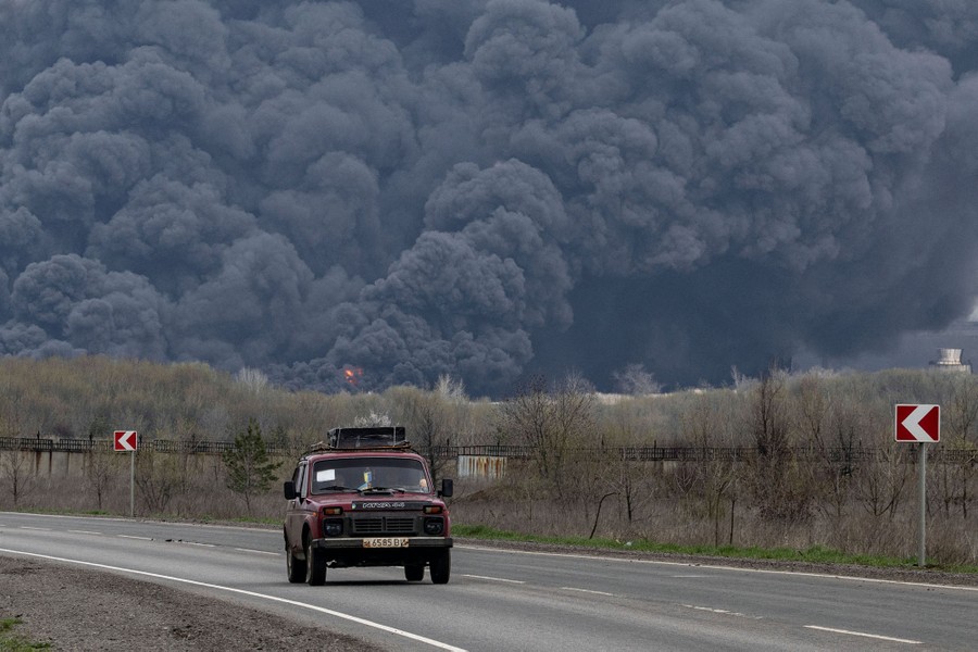 A car drives down a road; a wall of black smoke rises behind it in the distance.