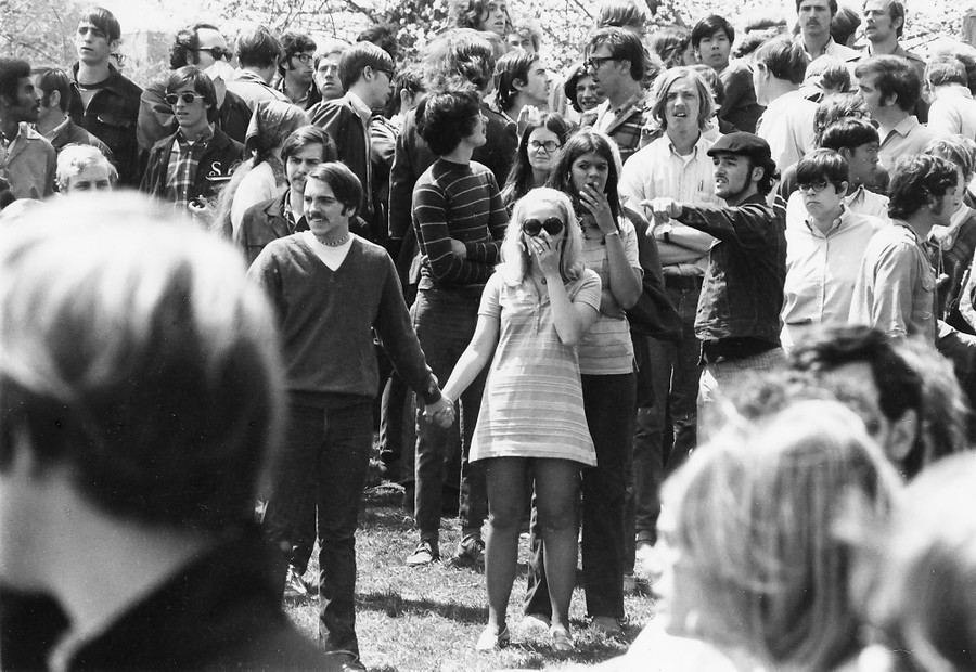 A group of protesters stand on a hillside, looking on, some with their hand over their mouth.