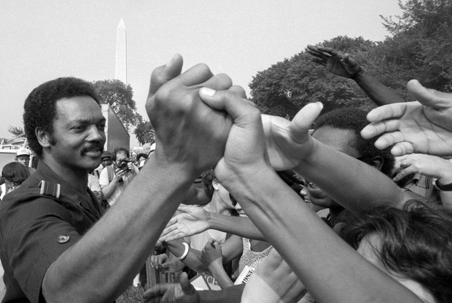 A black and white photograph of Jesse Jackson clasping hands with members of a crowd, with the Washington Monument in the background