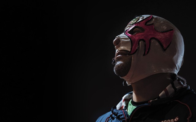 A color photograph of a man wearing a skin-tight white-and-pink luchador mask, set against a black background.