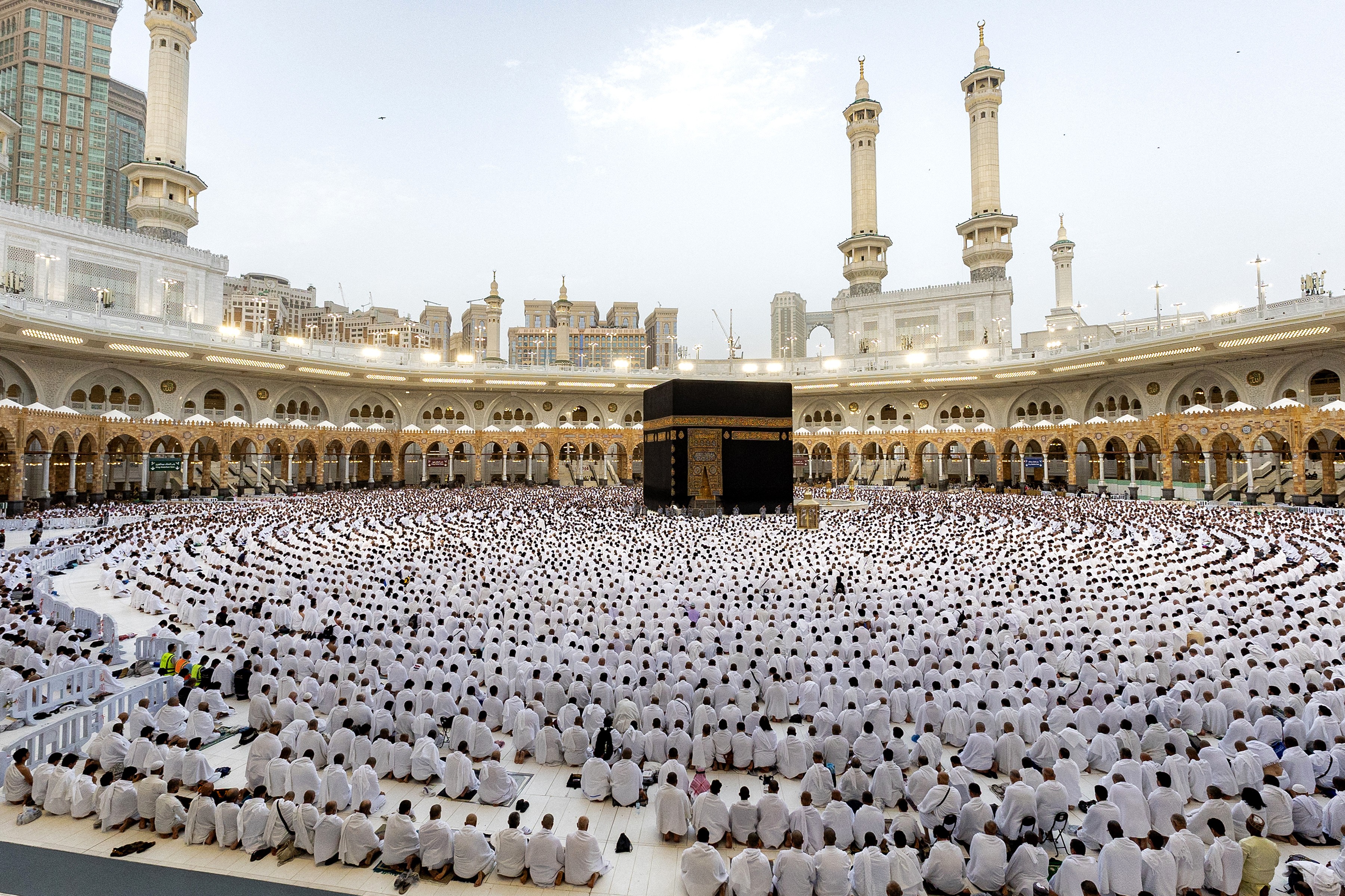 A large group of worshipers sit in concentric circles around a large cube shrouded with black fabric.