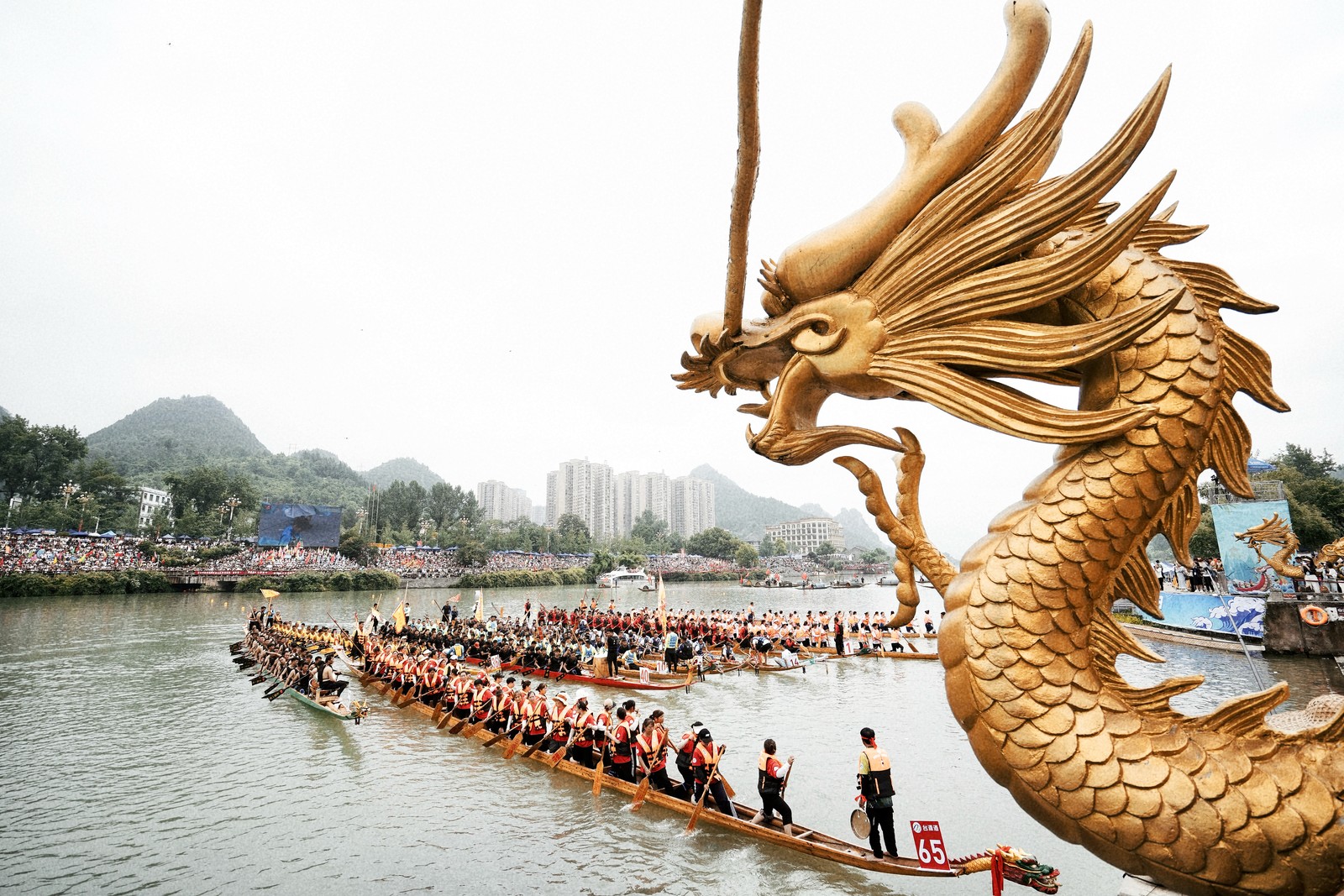 Racers stand in several very long and narrow boats, waiting for a race, beneath an elaborate carved dragon head.