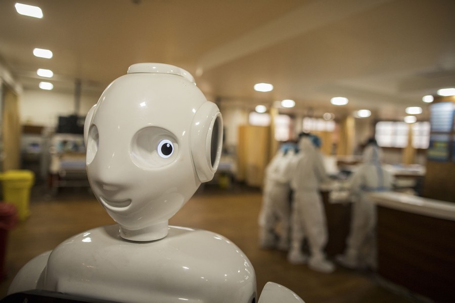 The face of a humanoid robot is seen in a hospital, with medical workers in the background.