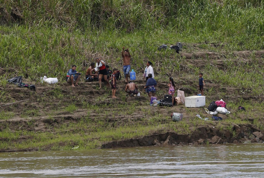 A small group of people with some belongings stand and sit on a river shore.