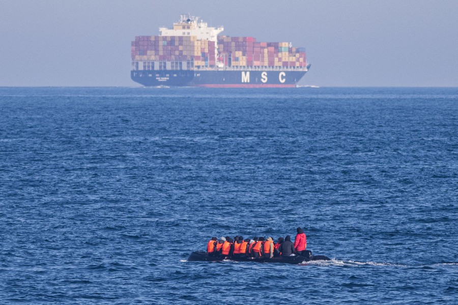 A small inflatable boat is crowded with people in a body of water; a large cargo ship sits on the horizon.