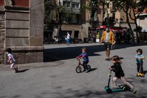 A man in a mask oversees a group of children playing on bikes and scooters in a plaza outside.