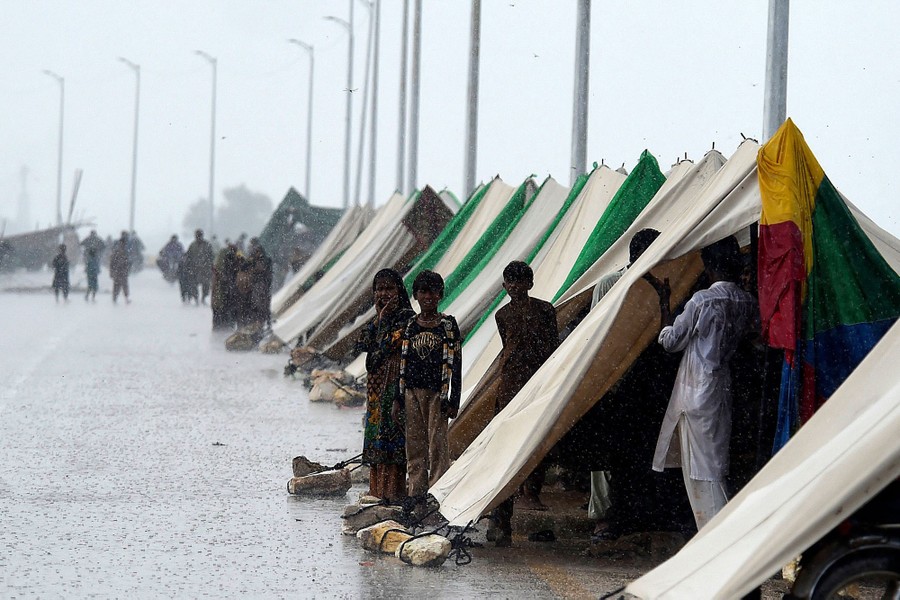 People stand in and near a line of tents set up alongside a road in heavy rain.