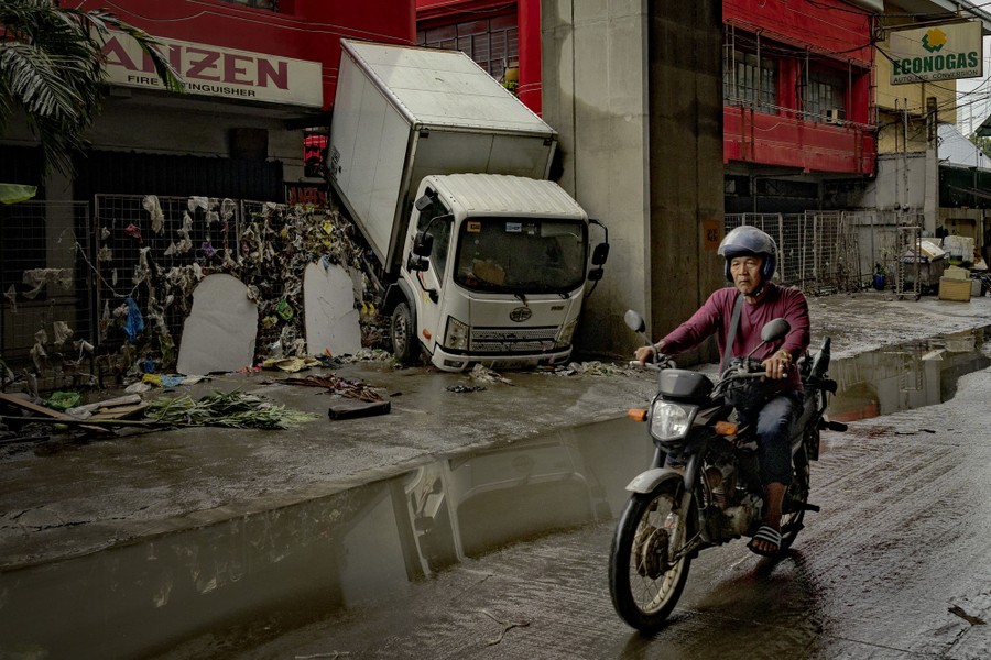 A person rides a motorcycle past a storefront where a small delivery truck has been pushed up and over a fence by flooding that has now receded.