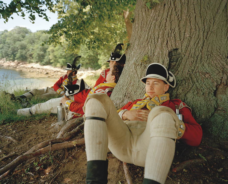 photo of several reenactors dressed as British soldiers sitting in the shade on grass and leaning against the base of a very large tree during a pause, with the shore in background