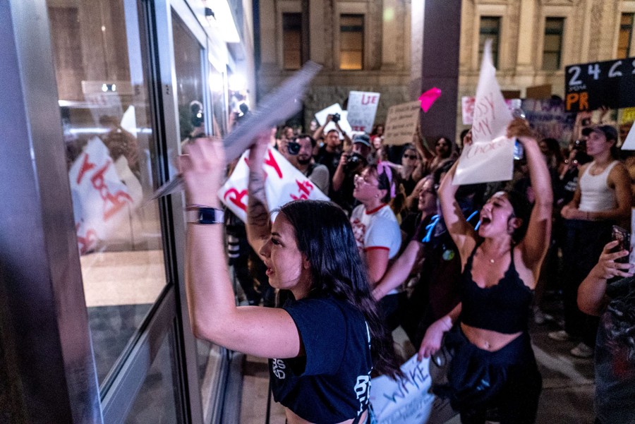 Protesters chant and hold up signs outside a building's entrance.