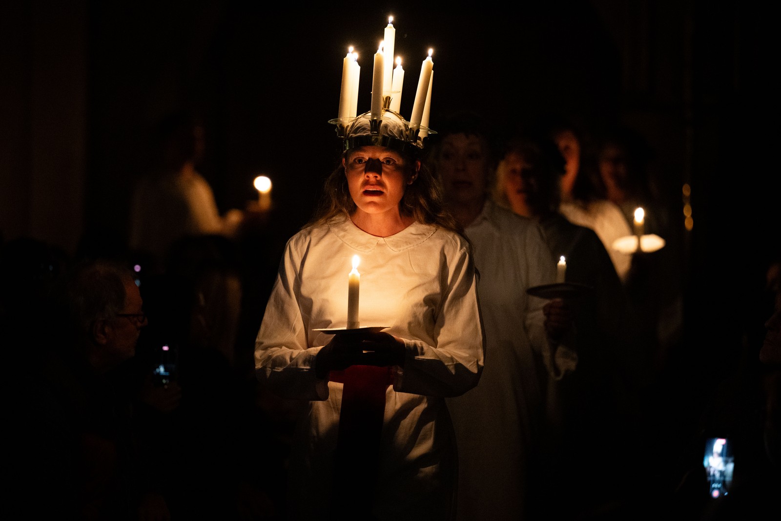 People carry candles during a choir performance, with the person at center wearing a crown of lit candles.