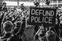 Fotografía en blanco y negro de una multitud que protesta con los puños en alto y un cartel que dice, “DESFINANCIAR LA POLICÍA”.