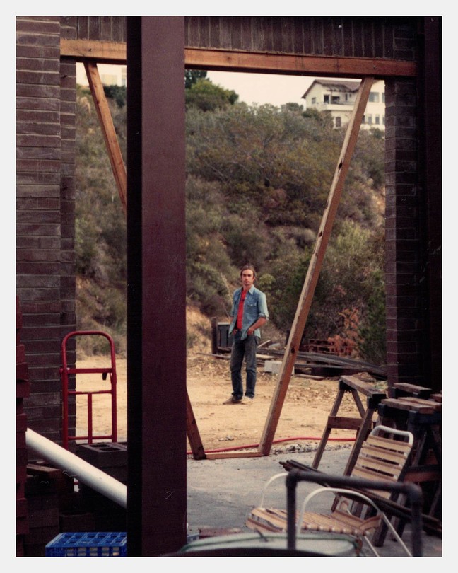 A color photograph of a man standing outside, seen from inside of a garage.