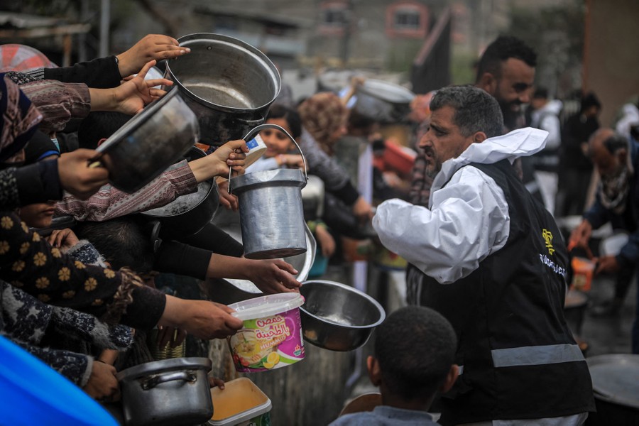 A crowd of Palestinians reach out their hands, holding pots and buckets for food at a donation point.