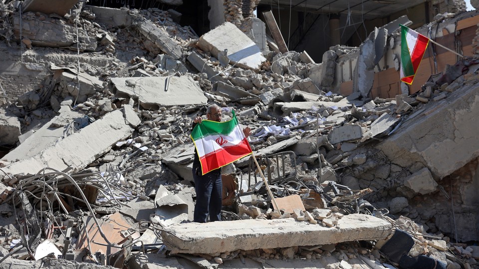 Photograph of a man holding up an Iranian flag atop the wreckage of a bombed building