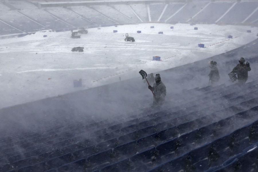 People holding snow shovels work in the stands of a stadium as wind-whipped snow swirls around.