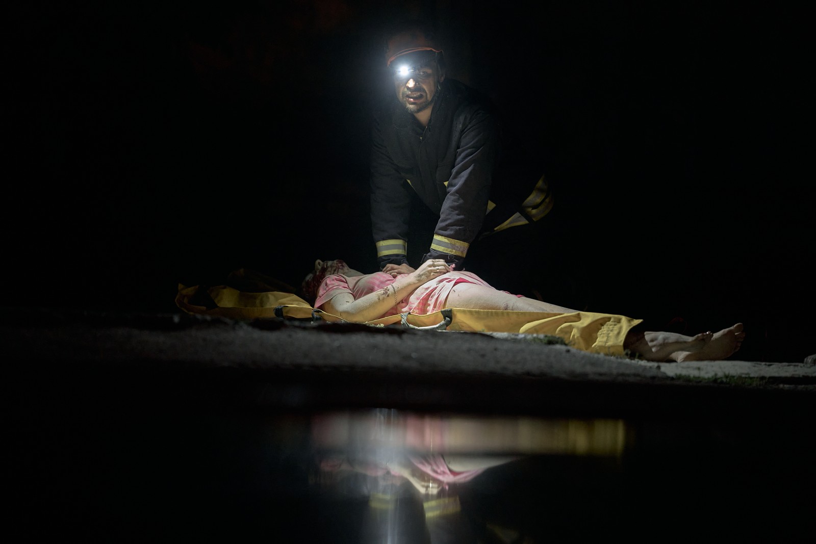 A rescue worker kneels beside an injured person on the ground, outside a damaged building at night.