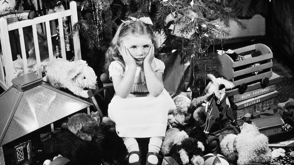 A girl sits with her face in her hands, looking disgruntled, surrounded by gifts under a Christmas tree